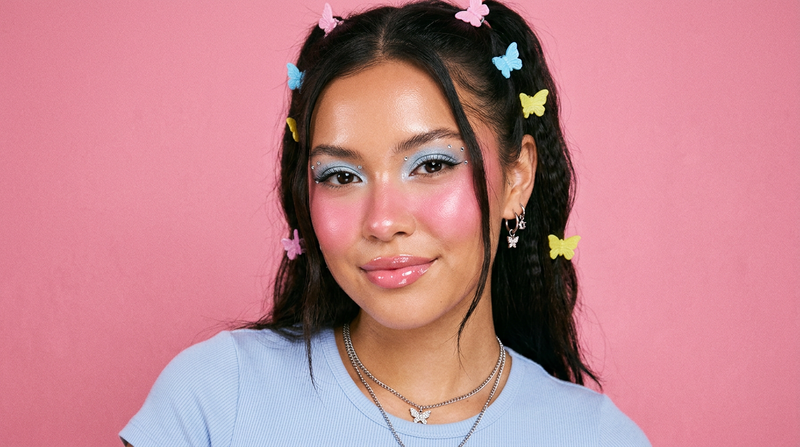 Model with flushed glowing pink cheeks Y2K makeup look using Satin Blush Powder, rhinestone accessories, butterfly hair clips, bubblegum pink background